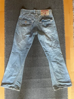 Ljusblå True Religion jeans bootcut - Ljusblå jeans från True Religion med bootcut passform. Jeansen har klassiska kontrastsömmar, broderade bakfickor med knapp samt en liten slitning på ena benet framtill.