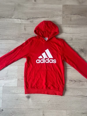 Röd hoodie från Adidas med logga - Snygg röd hoodie från Adidas med stor vit logga på bröstet. Klassisk känguruficka framtill och huva. Tillverkad i mjukt material, perfekt för en avslappnad stil. Passar dig som gillar sportiga och bekväma plagg.