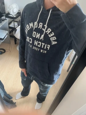 Mörkblå hoodie Abercrombie & Fitch - Snygg mörkblå hoodie från Abercrombie & Fitch med vit text framtill och klassisk känguruficka. Huvan har vita dragsnören och tröjan är gjord i mjukt bomullsmaterial. Perfekt för en avslappnad och trendig look.