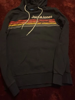 Jack & Jones svart hoodie med ränder - Snygg svart hoodie från Jack & Jones med färgglada ränder i rött, orange, gult och vitt över bröstet. Trycket 'Jack & Jones Originals' framtill och vita dragsnören i huvan. Perfekt för en avslappnad streetwear-look.