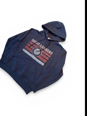 Mörkblå hoodie Saint Laurent - Mörkblå hoodie från Saint Laurent med tryck i vitt och rött framtill. Klassisk känguruficka och justerbar huva med snören. Hoodien har en avslappnad passform och är tillverkad i mjukt bomullsmaterial. Perfekt för en casual och trendig look.