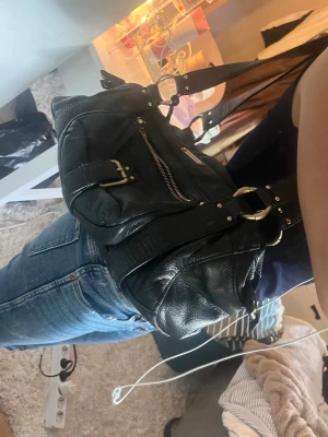Svart axelremsväska i skinn - Gammal micheal kors väska. Snygg svart handväska i mjukt skinn med axelrem och detaljer i guld. Väskan har dragkedja framtill, dekorativa spännen och flera fack. Perfekt storlek för det viktigaste och passar till de flesta outfits.