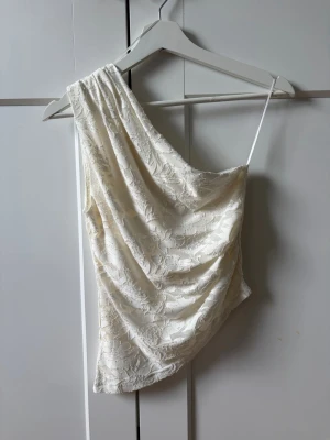 Vit spets topp oneshoulder - Säljer en vit oneshoulder topp med tunna axelband och vacker spetsdetalj. Toppen har en asymmetrisk design och är perfekt för dig som vill sticka ut lite extra. Materialet är mjukt och följsamt med blommigt spetsmönster.