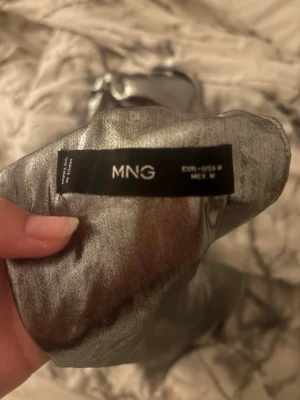 Silver axelbandslös topp från MNG - Säljer en glittrig silverfärgad axelbandslös topp från MNG. Toppen har en lös passform och är tillverkad i ett skimrande syntetmaterial som verkligen sticker ut. Perfekt för dig som vill ha något extra till din outfit.