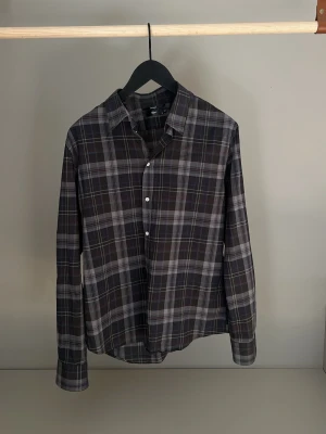 Tunn mörkgrå rutig skjorta från Filippa K - Snygg mörkgrå skjorta från Filippa K med rutigt mönster i vitt och grått. Skjortan har klassisk krage, långa ärmar och knappar framtill. Väldigt tunt tyg vilket passar bra både för sommaren eller att ha under en tröja. Perfekt för dig som gillar en avslappnad men stilren look.