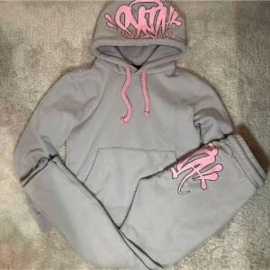 Grått loungewear-set med rosa detaljer - Säljer ett grått loungewear-set med hoodie och byxor. Hoodien har rosa snören och rosa graffiti-tryck på huvan, samt en stor magficka. Byxorna har matchande rosa tryck på benet och resår i midjan. Perfekt för chill dagar hemma.