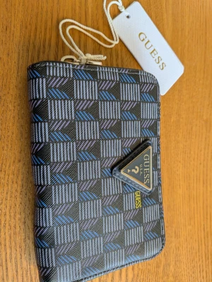 Mönstrad plånbok från Guess - Snygg plånbok från Guess med svart botten och geometriskt mönster i blått och grått. Plånboken har dragkedja i guld, flera fack för kort och myntfack med dragkedja. Guess-logga i metall på framsidan. Tillverkad i konstläder.