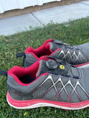 Grå och röda stålhettaskor med BOA-snörning - Säljer ett par grå stålhellaskor med röda detaljer och vit sula. Skorna har ett smart BOA-snörningssystem och mönstrade sidopaneler i vitt och rött. Insidan är fodrad i rött tyg och sulan är stötdämpande. Perfekta för dig som vill ha både stil och funktion.