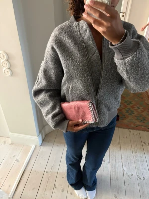 Rosa handväska med kedjedetaljer - Säljer en liten rosa handväska i skinnimitation med silverfärgade kedjedetaljer runt kanterna. Väskan har en rektangulär form och dragkedja upptill. Perfekt för dig som vill ha en cool accessoar som sticker ut. Kommer även med en kedja men kedjan är trasig 