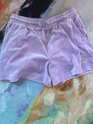 Ljuslila velourshorts Juicy Couture - Säljer ett par ljuslila velourshorts från Juicy Couture med snörning i midjan och fickor på sidorna. Shortsen har en glittrig JC-logga i strass på ena benet och är perfekta för en avslappnad och trendig look.