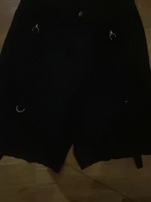 Svarta shorts med ringdetaljer Divided - Den andra bilden är i blixt! Dem är svarta! Dem ser bara konstiga ut med blixt på! Säljer ett par svarta shorts från Divided H&M med flera silverfärgade ringdetaljer framtill. Shortsen har hög midja och knappstängning. Perfekta för dig som gillar en edgy stil.