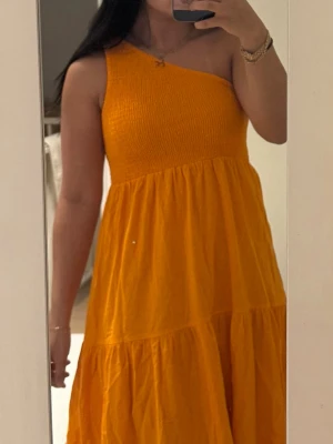 Orange axelbandslös sommarklänning - Snygg orange klänning med en axel, smockad överdel och volangkjol. Perfekt för varma dagar och ger en trendig look. Tillverkad i ett lätt och luftigt tyg som känns bekvämt hela dagen. Passar dig som vill sticka ut med färg och stil.