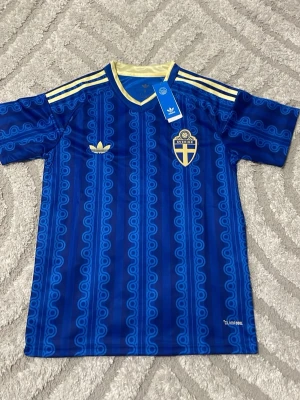 Blå Sverige tröja från Adidas - Säljer en blå Sverige fotbollströja från Adidas med retroinspirerat mönster, gula detaljer och broderad svensk landslagssköld. Tröjan har korta ärmar, rund halsringning och tre gula ränder på axlarna. Tillverkad i lätt funktionsmaterial.