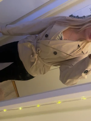 Beige trenchcoat med knytskärp - Stilren beige trenchcoat med dubbelknäppning och knytskärp i midjan. Jackan har klassisk krage, svarta knappar och är tillverkad i ett lätt tyg som passar perfekt till vår och höst. Snygg och tidlös modell som funkar till många outfits.