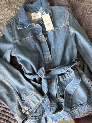 Ljusblå jeansjacka med knytskärp - Trendig ljusblå jeansjacka med knytskärp i midjan och stora fickor framtill. Klassisk krage och kontrastsömmar ger en snygg look. Perfekt för dig som vill ha en avslappnad men stilsäker outfit.