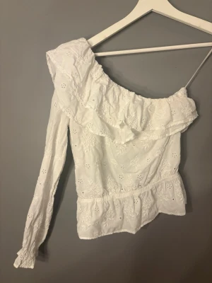 Vit oneshoulder topp med volang - Säljer en vit oneshoulder topp med broderade detaljer och volang längs halsringningen. Toppen har en lång ärm och är tillverkad i ett lätt bomullstyg med fina hålbroderier. Perfekt för dig som vill ha något unikt och trendigt! 