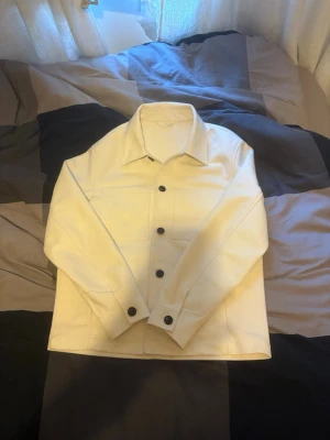 Vit overshirt med svarta knappar - Stilren vit overshirt med raka linjer och stora bröstfickor. Jackan har klassisk krage och svarta knappar framtill som kontrast. Materialet ser ut att vara tjockt och mjukt, perfekt för lager på lager. Enkel och modern design som passar till mycket.
