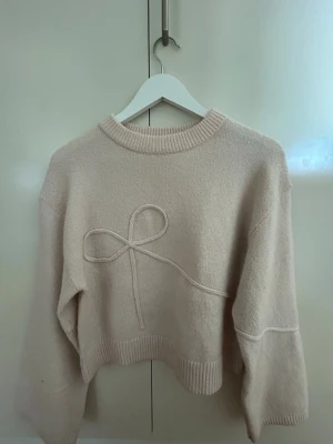 Beige stickad tröja med rosett - Söt ljusrosa stickad tröja med en dekorativ rosett framtill. Tröjan har en avslappnad passform, ribbade muddar vid ärmslut och nederkant samt rund halsringning.