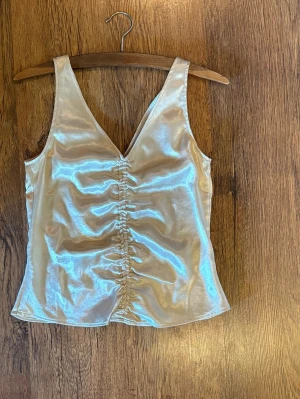 Champagnefärgad satinlinnetopp - Snygg champagnefärgad linnetopp i satin med v-ringning och rynkdetalj framtill. Toppen är ärmlös och har en figurnära passform.