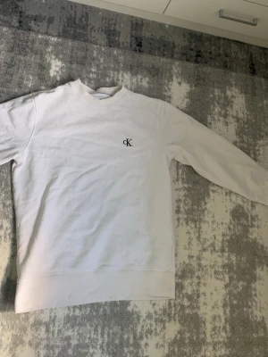 Vit sweatshirt från Calvin Klein - Vit sweatshirt med rund hals och diskret CK-logga broderad på bröstet. Tröjan har långa ärmar och ribbade muddar vid ärmslut och nederkant. Perfekt för en clean och stilren look.