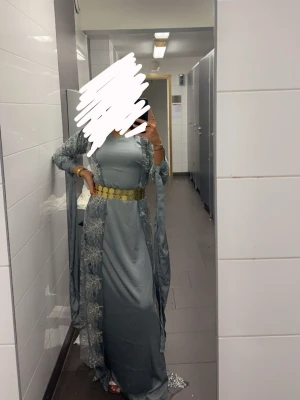 Lång grå klänning med spetsdetaljer - Elegant långklänning i grått med vackra spetsdetaljer och ett brett guldigt bälte i midjan. Klänningen har lång ärm och ett transparent spetslager ovanpå, vilket ger en lyxig och unik look. Perfekt för dig som vill sticka ut.