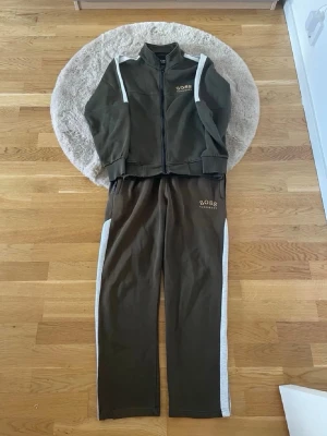 Grön BOSS set - Snyggt set från BOSS i mörkgrönt med vita ränder längs ärmar och ben. Setet består av en ziptröja med hög krage och matchande byxor med resår i midjan. BOSS-logga i vitt på både tröja och byxa. Perfekt för chill dagar hemma.