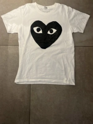 Vit Comme des Garçons Play t-shirt - Vit t-shirt från Comme des Garçons Play med det ikoniska svarta hjärtat med ögon tryckt på bröstet. Klassisk rund hals och korta ärmar. Perfekt för dig som gillar streetwear och vill sticka ut med en unik design.