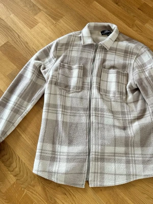 Beige rutig overshirt Hampton Republic - Overshirt i mjukt beige och vitt rutigt tyg från Hampton Republic. Två bröstfickor, dragkedja framtill och klassisk krage. Perfekt lager-på-lager-plagg med avslappnad passform och stilren look.