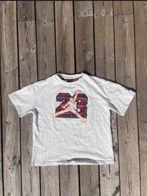 Vit t-shirt med tryck av 23 och Jumpman - Vit t-shirt med stort tryck av siffran 23 i röd och svart rutig design samt en orange kontur av Jumpman-loggan på bröstet. Klassisk rund hals och korta ärmar. Perfekt för dig som gillar sportig streetstyle.