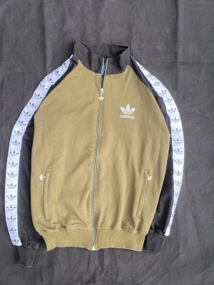 Olivgrön Adidas track jacket - Snygg olivgrön track jacket från Adidas med dragkedja framtill och vita Adidas-loggor längs ärmarna. Svarta detaljer på krage och ärmar samt två fickor med dragkedja. Perfekt för sport eller streetwear.