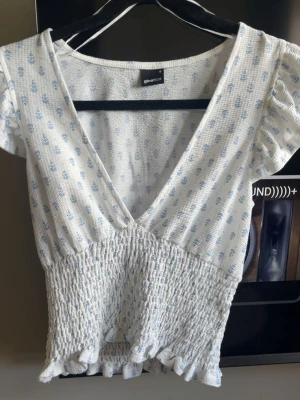 Vit blommig omlottopp från Gina Tricot - Supersöt vit topp med små blå blommor från Gina Tricot. Omlottmodell med v-ringning, korta puffärmar och smockad midja som ger en snygg passform. Volangkant nedtill för extra detalj. Perfekt för sommarens alla tillfällen!