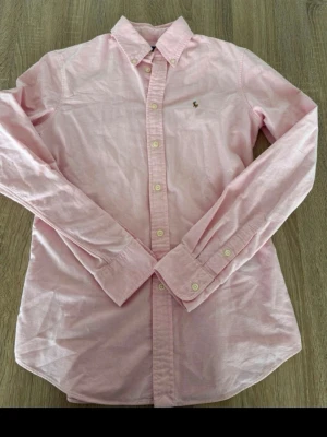  Polo Ralph Lauren Skjorta - Klassisk ljusrosa skjorta från Polo Ralph Lauren med broderad logga på bröstet. Skjortan har långa ärmar, knappar framtill och är tillverkad i mjuk bomull. Perfekt för en stilren och avslappnad look. 