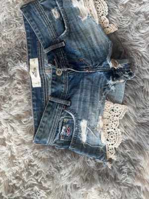 Blå jeansshorts med spets från Hollister - Säljer ett par blå jeansshorts från Hollister med slitna detaljer och snygg spetskant nedtill. Shortsen har klassisk femficksmodell, knappgylf och råa kanter. Perfekta för sommaren och ger en avslappnad, trendig look.