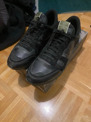 Svarta sneakers från Valentino - Säljer ett par svarta sneakers från Valentino med coola camo-detaljer och mix av mocka och läder. Skorna har svarta snören, låg profil och en chunky sula med nitar på hälen. Perfekta för dig som gillar streetstyle och exklusiva märken.