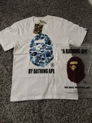 Vit BAPE t-shirt med blått camotryck - Säljer en vit t-shirt från A Bathing Ape med det ikoniska apansiktet i blått camomönster på bröstet och texten 'BY BATHING APE' under. Klassisk passform och rund hals. Perfekt för dig som gillar streetwear och vill sticka ut.