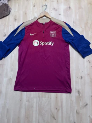 Barcelona träningströja Nike - Säljer en Barcelona träningströja från Nike i vinrött med blå ärmar och beige axelparti. Tröjan har halv dragkedja, klubbmärke och Spotify-logga på bröstet. Perfekt för fotbollsträning eller som supporterplagg.