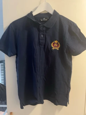 Mörkblå pikétröja med brodyr - Klassisk mörkblå pikétröja med kort ärm och krage. Tröjan har knappar framtill och en broderad detalj med emblem på bröstet. Perfekt för en stilren och sportig look.