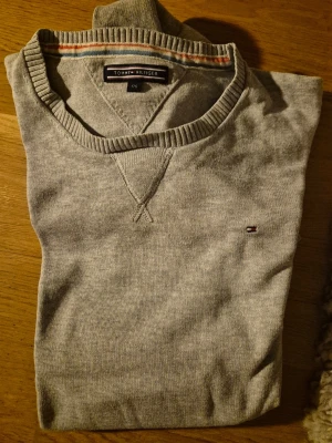 Grå tröja från Tommy Hilfiger - Snygg grå tröja från Tommy Hilfiger med rund halsringning och ribbade muddar. Liten broderad logga på bröstet och diskreta ränder på insidan av kragen. Perfekt för en clean och stilren look.