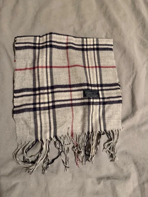 Burberry halsduk grå  - Snygg grå halsduk med klassiskt rutmönster i marinblått, vitt och rött. Halsduken har fransar i båda ändar och en mjuk känsla. Perfekt accessoar för att lyfta din outfit med en tidlös look.