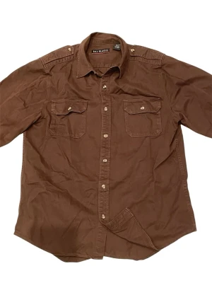 Brun vintage-skjorta från Bill Blass - Säljer en brun skjorta med två bröstfickor och snygga knappar framtill. Skjortan har axelklaffar och klassisk krage, samt långa ärmar. Perfekt för dig som gillar en enkel men ändå stilren look.