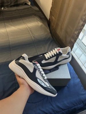 Mörk blå Prada cups  - Snygga sneakers i svart och vitt med glansiga partier och meshdetaljer på sidorna. Skorna har vita snören och en vit sula som ger en fräsch look. Perfekta för dig som gillar stilrena och moderna sneakers.