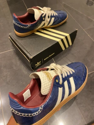 Adidas Wales bonner blå sneakers - Snygga Adidas sneakers i blått med krokodilpräglat mönster, vita detaljer och klassiska tre ränder på sidan. Skorna har vita snören, röd insida och gummisula. Tungan är vit med gulddetaljer och boxen medföljer.