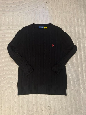 Svart kabelstickad tröja Polo Ralph Lauren - Kabelstickad svart tröja från Polo Ralph Lauren med rund halsringning och röd broderad logga på bröstet. Långa ärmar och ribbade muddar vid ärmslut och nederkant. Klassisk och stilren design som passar till mycket.