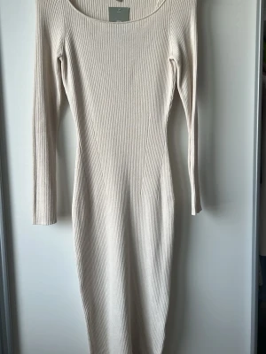 Beige ribbad långklänning - Snygg beige ribbad långklänning med lång ärm och rund halsringning. Klänningen har en kroppsnära passform och är tillverkad i ett mjukt, stretchigt material som passar perfekt till många olika stilar.