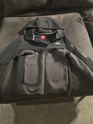 Svart Nike hoodie med dragkedja - Snygg svart hoodie från Nike med dragkedja framtill och huva. Har två stora fickor på magen och en liten ficka med dragkedja på ena ärmen. Nike-logga i grönt på bröstet. Tillverkad i mjukt material, perfekt för chill eller träning.
