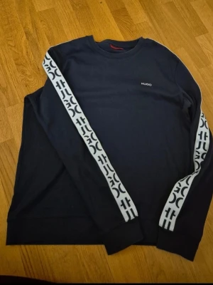Sweatshirt från Hugo Boss med logoband - Snygg svart sweatshirt från Hugo med vita logoband längs ärmarna och diskret Hugo-logga på bröstet. Tröjan har rund halsringning och är tillverkad i mjuk bomull. Perfekt för en avslappnad och trendig look.