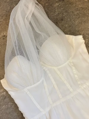 Vit långklänning med glittertopp - Supersnygg vit långklänning med glittrig mesh över bysten och axelbandslös design. Klänningen har en slits framtill och markerad midja för en elegant siluett. Perfekt för dig som vill sticka ut med en unik och glamorös look.