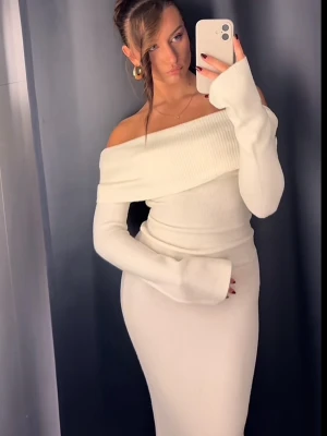 Vit offshoulder långklänning - Trendig vit långklänning med offshoulder-design och långa ärmar. Klänningen har en bred ribbad kant upptill och sitter figurnära för en snygg siluett. Perfekt för dig som vill ha en stilren och elegant look.