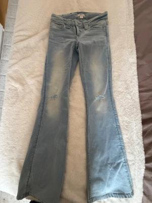 Ljusblå bootcut jeans med slitningar - Säljer ett par ljusblå bootcut jeans med slitningar på knäna och lätt tvättade detaljer. Jeansen har klassisk femficksdesign och utsvängda ben. Perfekta för dig som gillar trendigstil och vill ha ett par bekväma jeans med cool look. Använda väldigt fåtal gånger!💕
