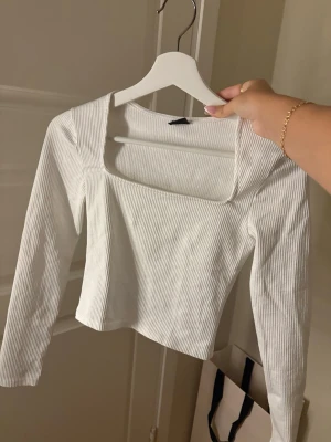Vit ribbad långärmad topp - Trendig vit ribbad topp med lång ärm och fyrkantig urringning. Croppad modell som passar perfekt till både jeans och kjol. 
